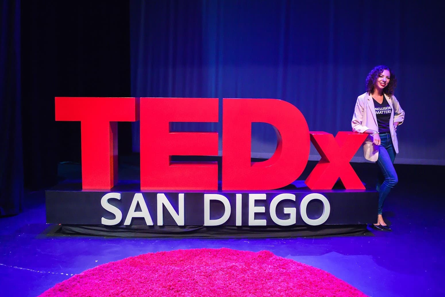 Woman in a blazer posing beside a large red TEDx San Diego sign on stage.