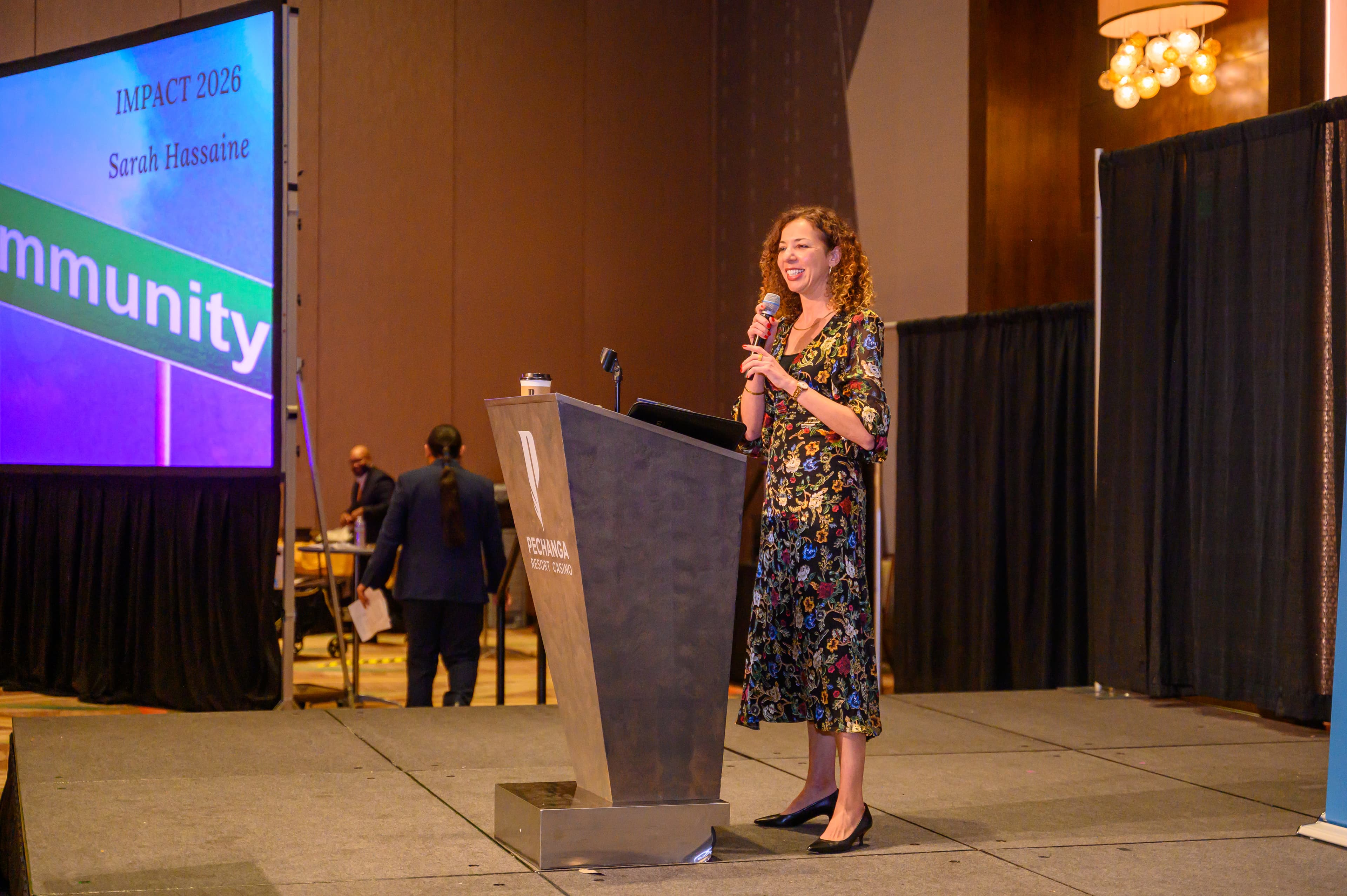 Woman in floral dress speaks at a podium during an Impact 2026 conference presentation.